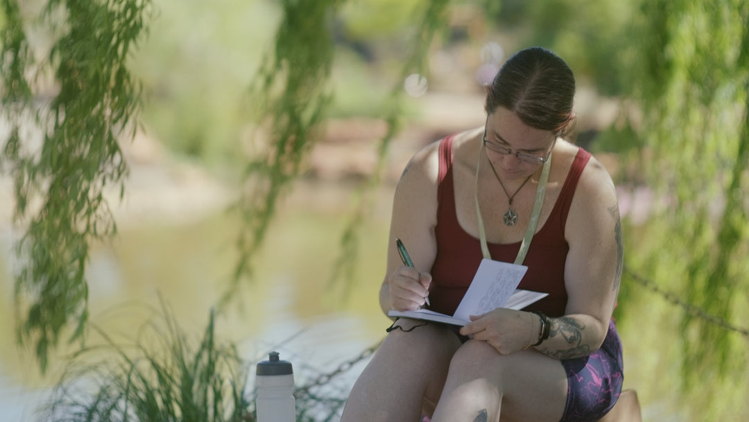 A front shot of a woman seating on a rock, with her head down as she writes in a journal. The tendrils of the leaves cascades at the back and an out of focus background of the lake.