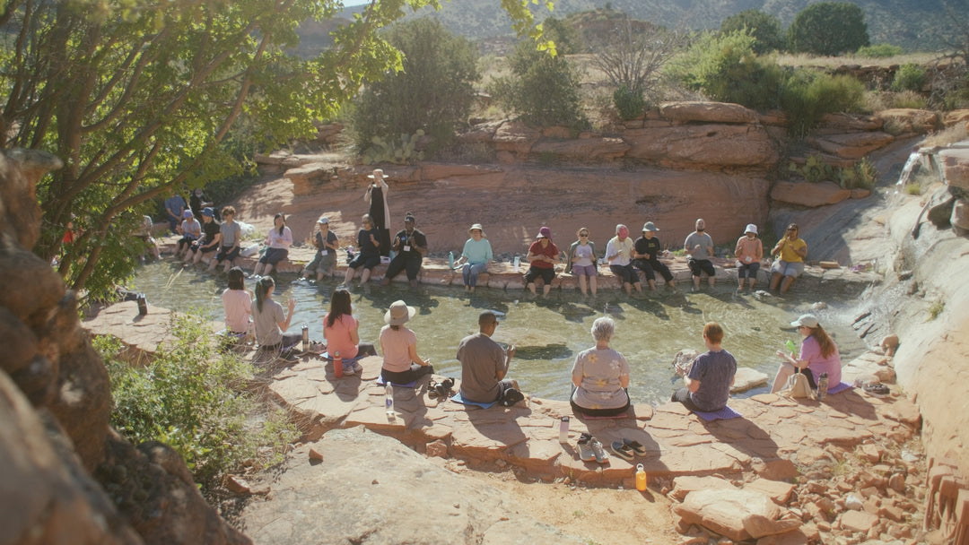 People seated surrounding a small pond lined with flat stones where  their feet dipped in it while surrounded by cliff rocks, few bushes and trees