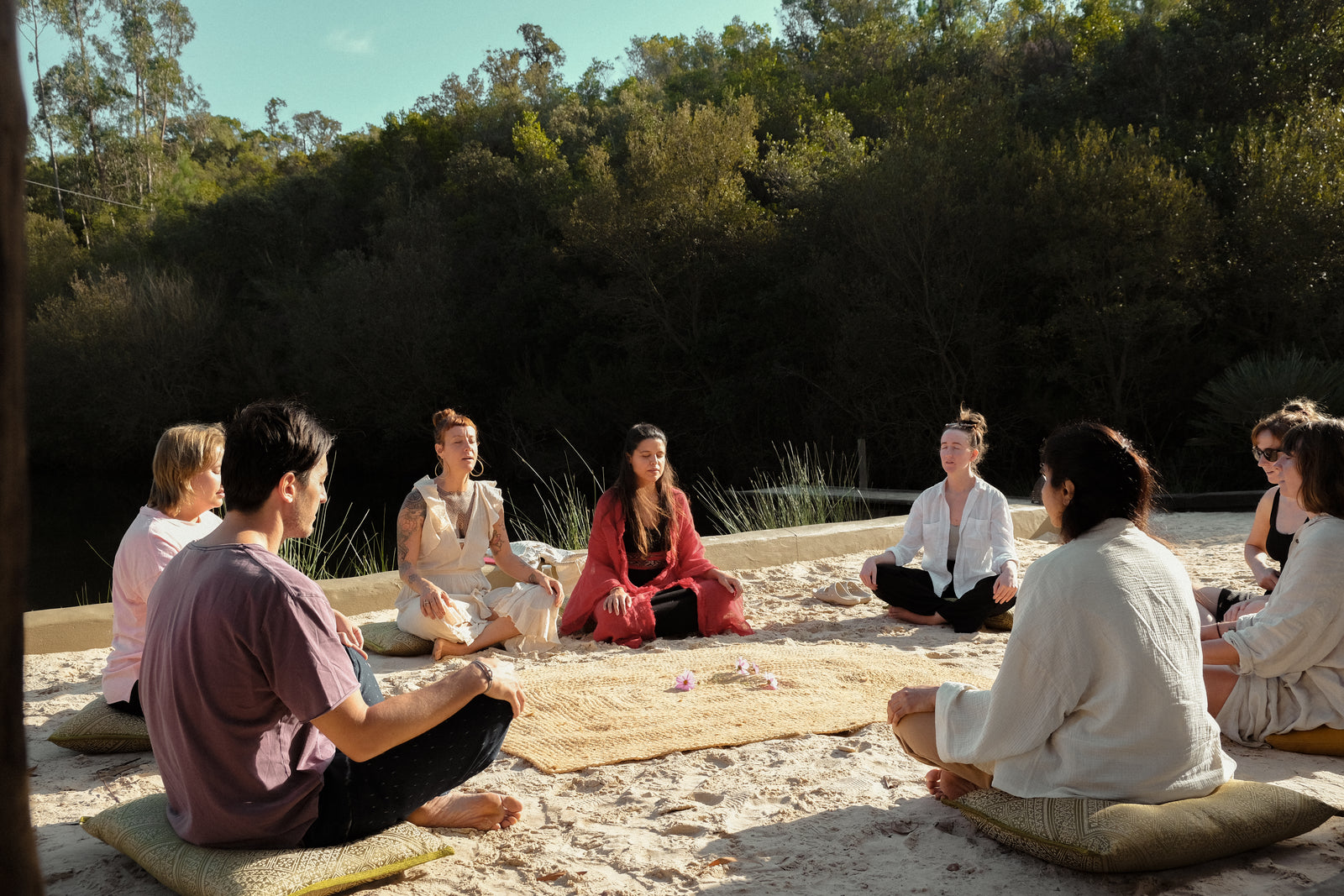 People outdoors, in circle formation, seated on a pillow on the sand, cross legged, hands on their knees, eyes closed and surrounded with trees.