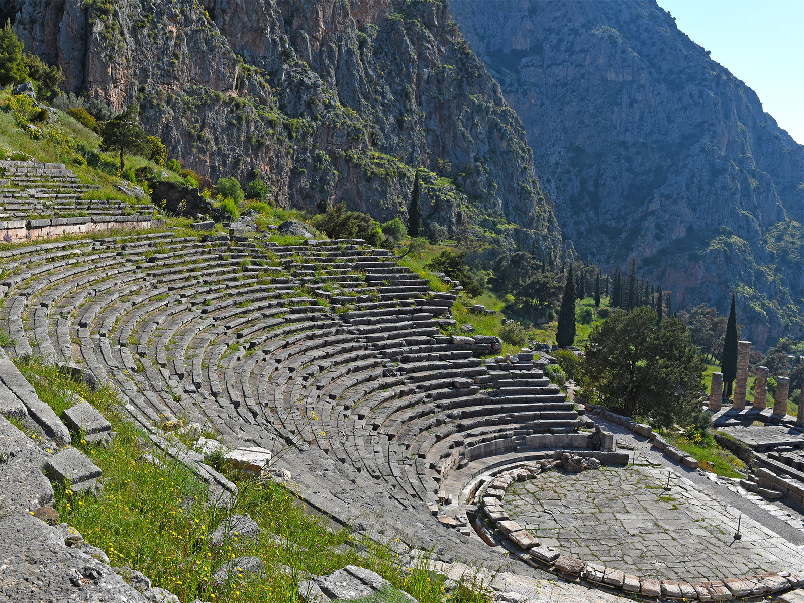 An ancient ampi-theater made of stones are installed in the side of a mountain surrounded with trees and mountain in the background
