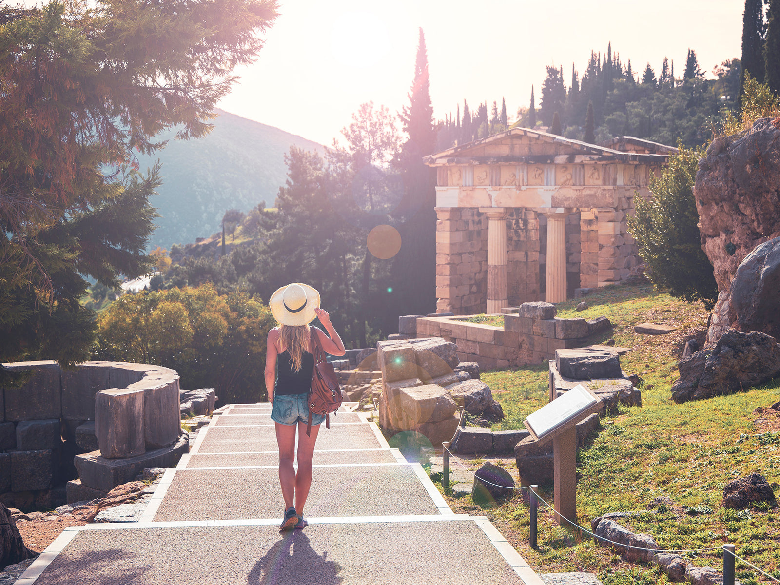 A backview of a woman standing, holding her hat, in the middle of a pathway with ruins of both sides and a small pantheon stands near, surrounded with trees and a mountain in the background