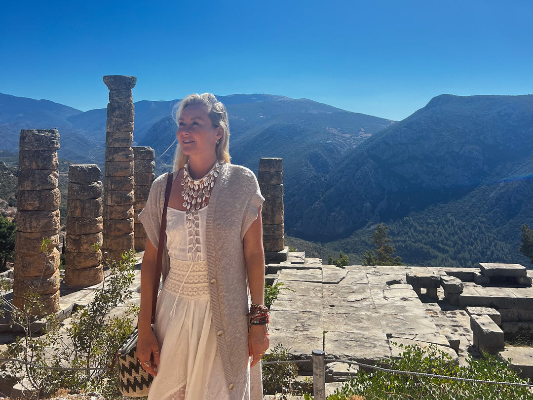 A woman standing forward facing, with a ruin at her back where a few columns that are stacked of stones and a mountain landscape in the background