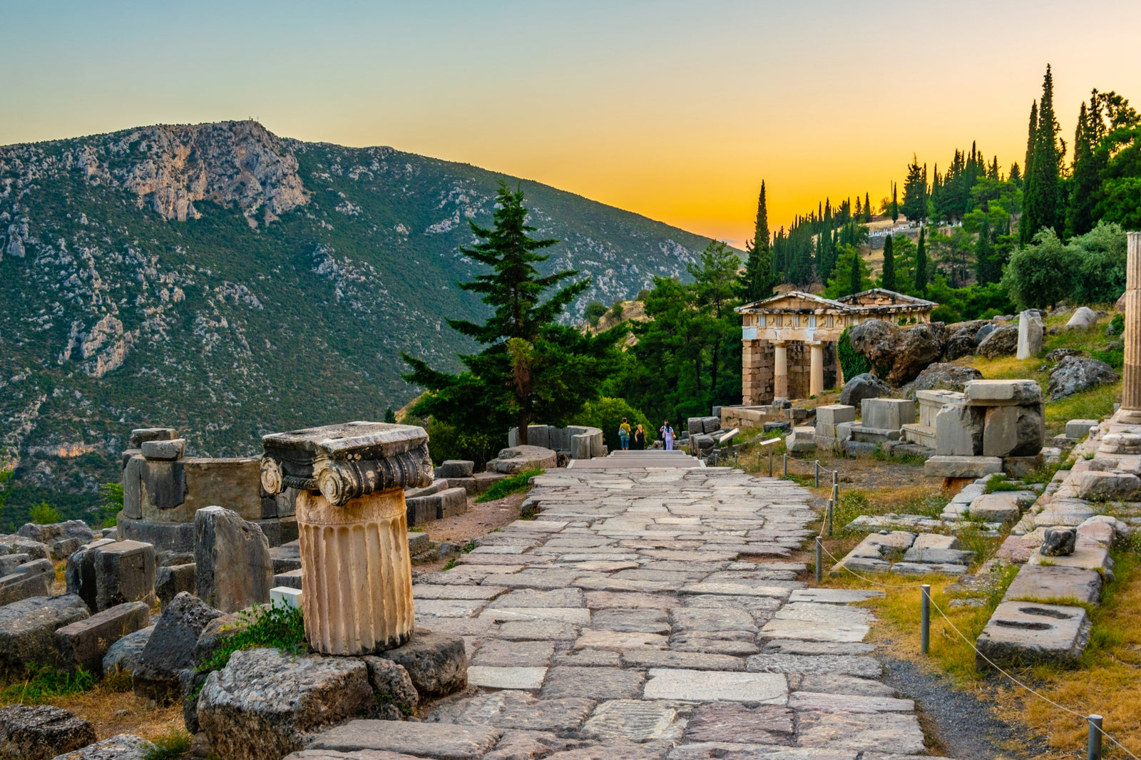 A pathway lain in stones with ruins on both sides and small pantheon that stands farther. surrounded with trees and a background of mountain.