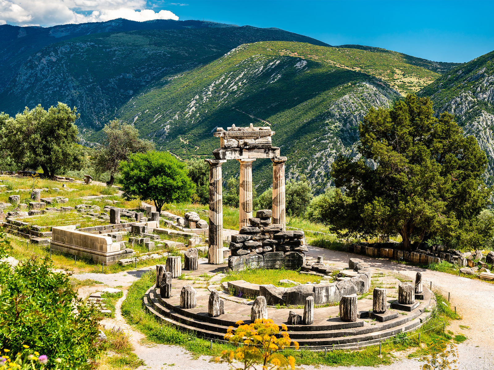 A ruin of a pantheon is in the middle where columns outside encircle  a ruined inside that's also in circle, where three complete columns stand. And other ruined structures behind it surrounded by trees. And a mountain landscape is on the background.