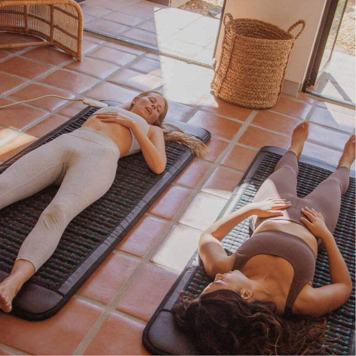 Two women lying besides each other, their heads are opposite from each other, on their mats in the floor and appears to be chatting.