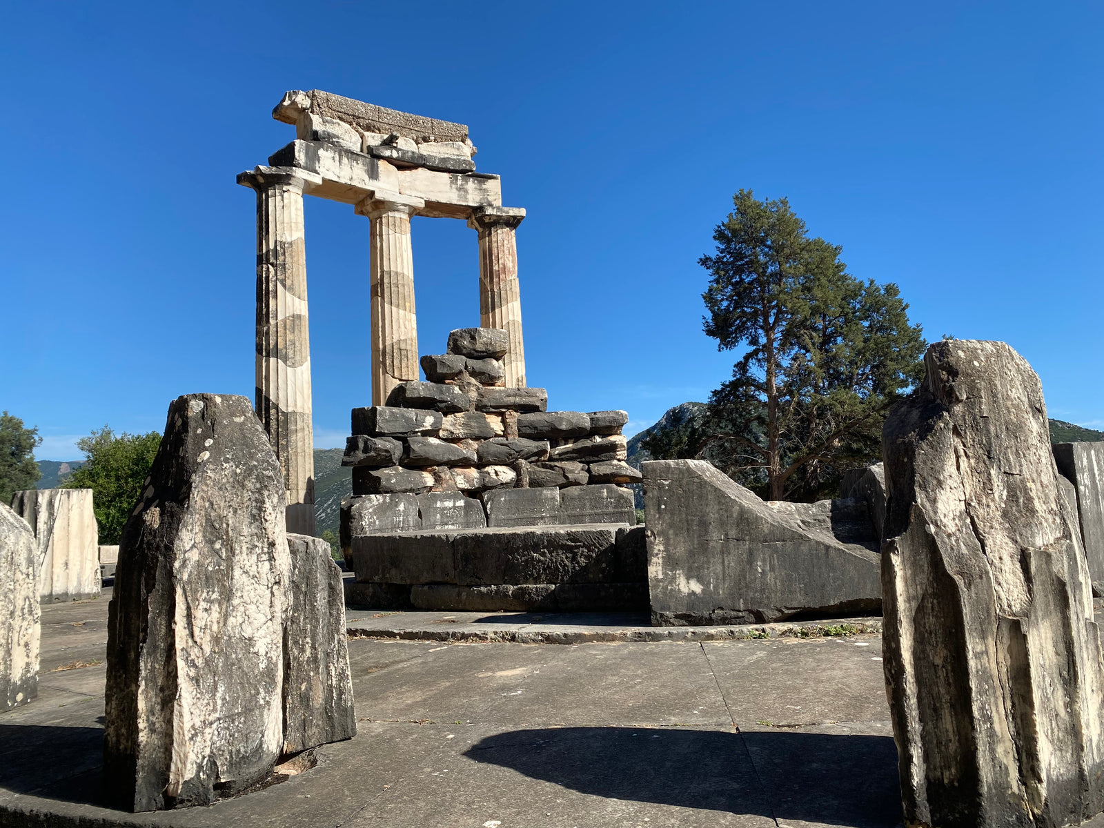 ruins of stones, standing and those stacked at the back with three columns and trees at the background