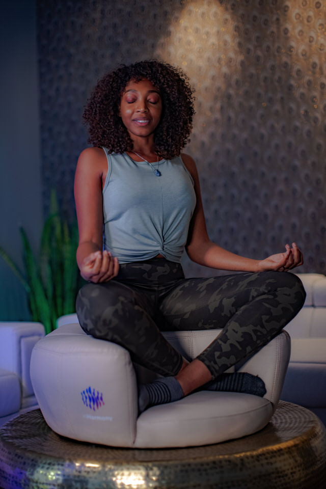 A woman sitting in a living room, atop a small cushion, in a elevated cross-legged manner, eyes closed, smiling and palms up on her knees.