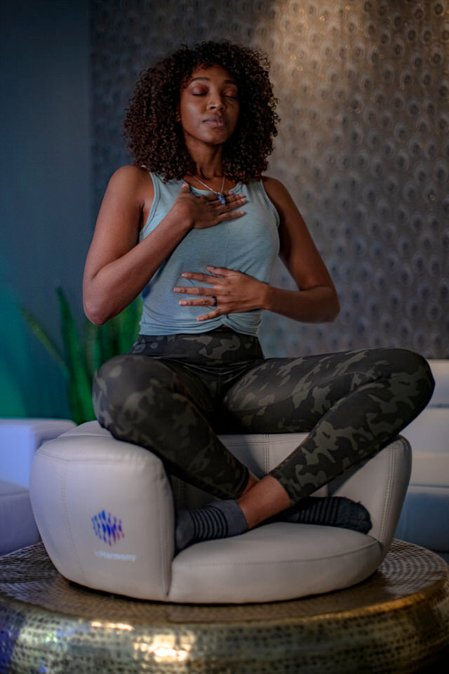 A woman in the middle of a room, sitting atop a white cushion, perched on a brass or golden-like low surface. She is cross legged, and her right hand on her chest and her left hand on her mid rib area, eyes closed, as if meditating