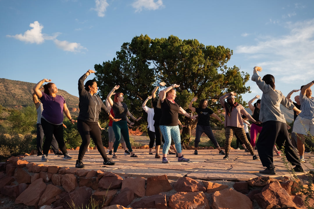 A mid-shot of people of their hands sideways over their head while facing the sun and their left foot stretched on a flat stoned floor surround by trees and a mountain at the background