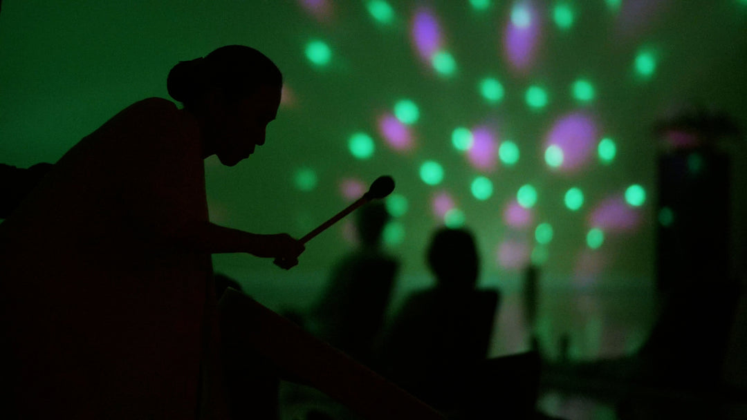 A side view of a silhouette of a woman with a drumstick on her hand. While an out of focus of silhouette of people sitting on the floor while a strobe of lights hitting the wall forming small and big circles.