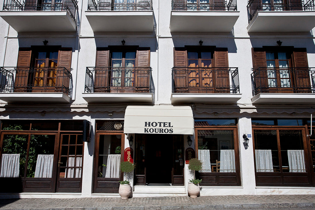 A building structure with a canopy in the entrance at the ground floor that says Hotel Kouros, the floor above showcase a steel balcony with slated doors