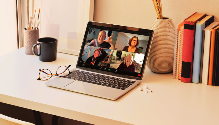 A desk set up with laptop on it showing 4 split screen with people in it. Glasses on left side, earphones on the right. Books, vase, cup and pencil cup in the back.