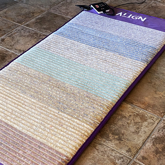 A flat lay of a mat on a floor. With crystals in between the horizontal stitchings with a text logo on the top that says Align and what looks like a remote and power contraption