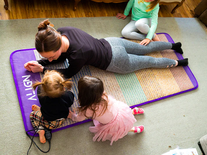 A woman is lying on a mat that is situated on the floor with 3 kids surrounding her seated