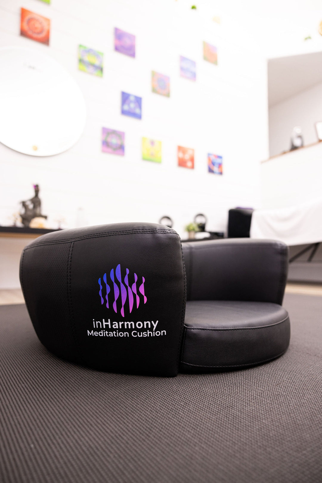 Sideview of a black cushion, with its logo highlighted on that has a squiggly lines and a text below inHarmony, Meditation Cushion, in the middle of a room indoors with a blurred background of a wall