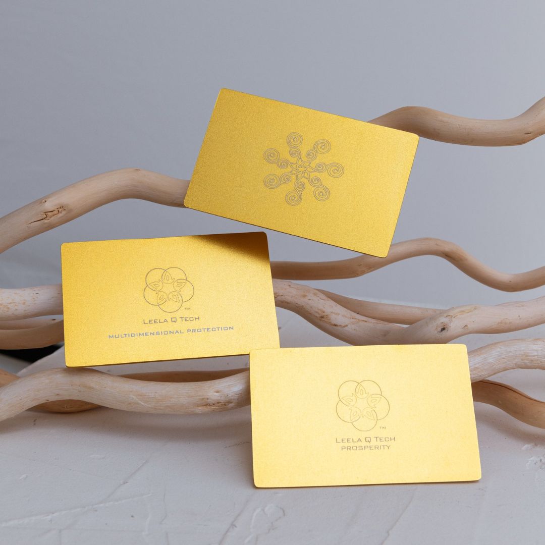 Threee golden yellow cards are perched on a light colored bark. The top card has a geometric logo of lotus-like flower in the middles and with seven spirals on each petal. In the middles is an interlocking symbol of five circles and a text below it that says Leela Q Tech, Multidimensional protection, and the below card has an interlocking symbol of five circles and a text below that says Leela Q Tech, Prosperity