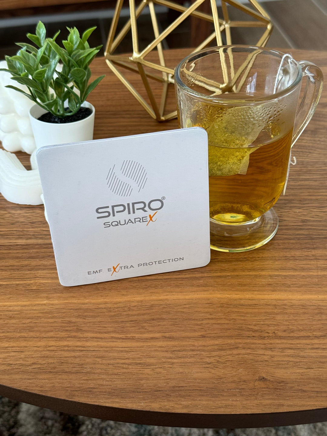 A square card with a text Spiro square x EMF extra protection stands next to a cup of tea, with a background of small pot of plant