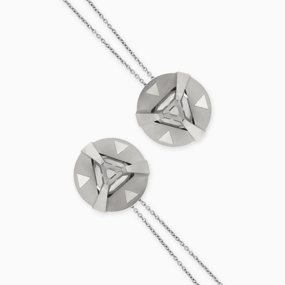 A close up and flat lay of two circular silver pendant in a necklace, side by side with the necklace opposite each other, have an encased crystal lodged in the outer pendant that has three prongs that houses it