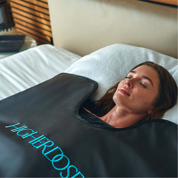 A woman lying on a bed, with her head on a pillow, eyes closed, inside an enclosed blanket with a text logo in the front that says Higherdose.