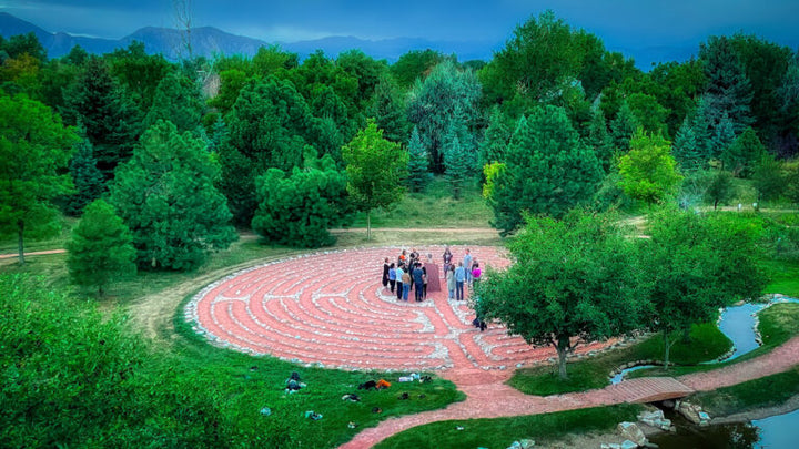 People gathering in the center of a flat labyrinth outlined by stones,  surrounded by trees