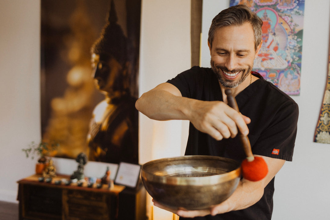 A man, smiling broadly, holding a golden bowl on his left hand, on his right, on top of the bowl, he holds small gong-like stick, the foam end touching the bowl. In the background is a wall with paintings and wall decorations.