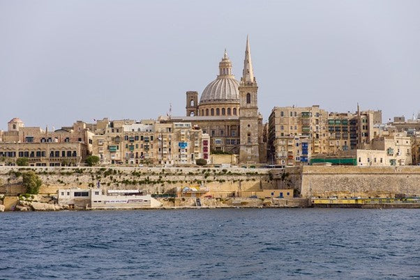 A body of water in the foreground, walls made of sandstone rocks line the coast. Further shows old looking buildings that looks like made of sandstone as well. An enormous cathedral is seen in between the building on a clear sky.