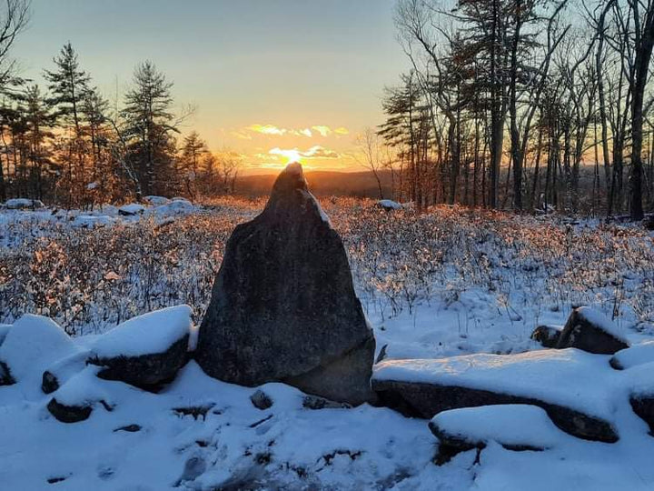 A landscape where rocks with snow are in the foreground and a triangular stone that covers the sun while the background is a field full of snow with many short weeds in it and big trees further and a long mountain even further almost covering the sun