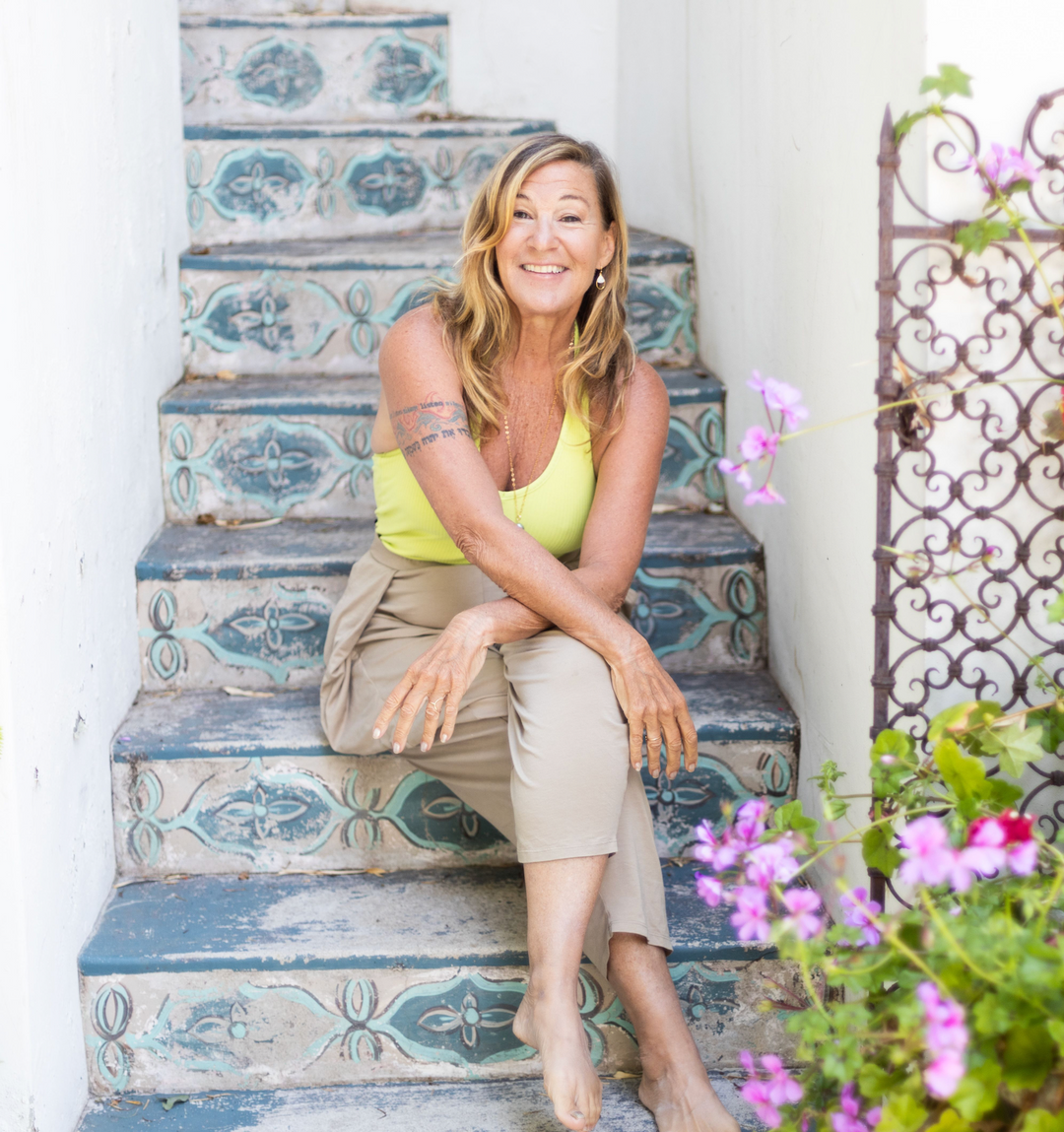 A woman barefoot, smiling, and sitting on the second bottom of a stairs, her legs crossed over and her hands lay across each other, relaxed on her knees.