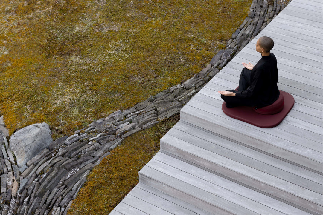 A bird's eyeview and sideview of person sitting crossed legged, palms on the knees facing upwards, on a cushion under a mat, perched on an outdoor deck. A stoned small pathway with short grasses in the front of the deck.