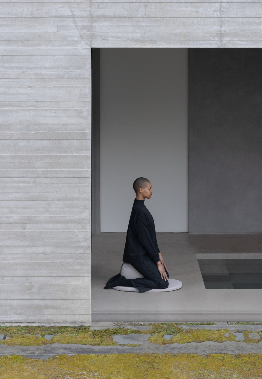 A person kneeling but seated on a cushion that is placed over a medium sized circle mat indoors but is much open to outdoors as it has an open wall with no doors