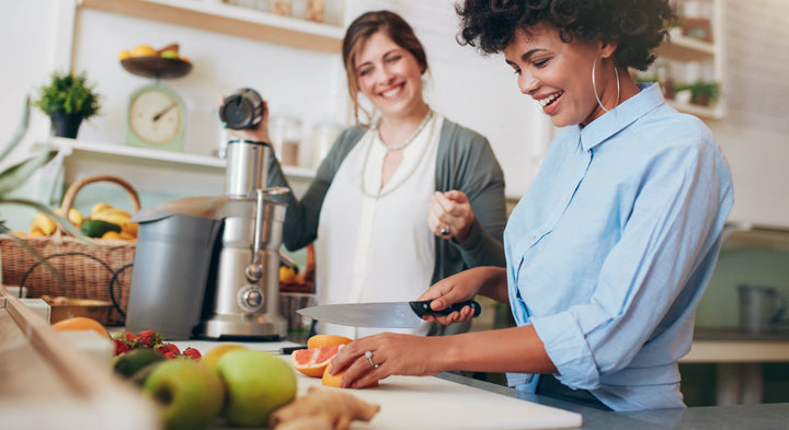 A set is in a kitchen, where one woman is cutting up a fruit, the other one is awaiting to put the fruits in the juicer