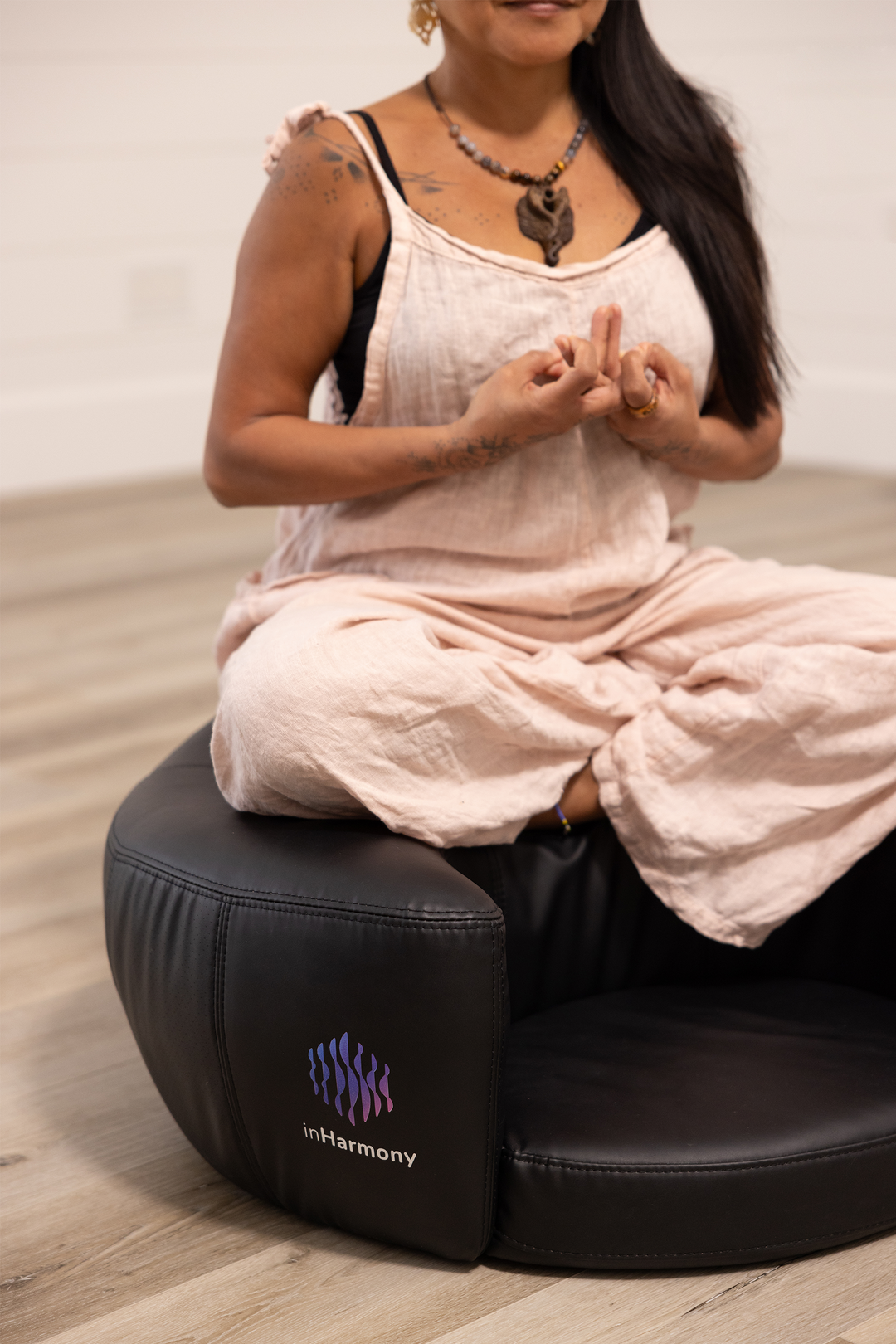 A picture of a woman, neck down, sitting cross-legged atop a black cushion indoors, her hands intertwined in her chest, as if meditating.