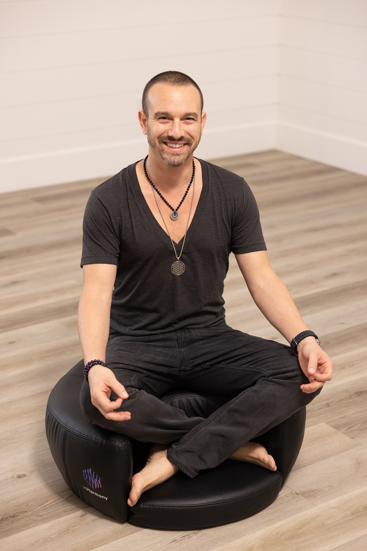 A man smiling sitting atop a cushion, cross legged, and hands on the knees with his index and thumb touching together