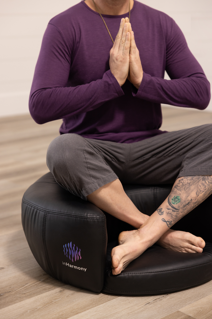 A mid-shot of a person, sitting atop a cushion, legs crossed, and his palms touching together in the chest