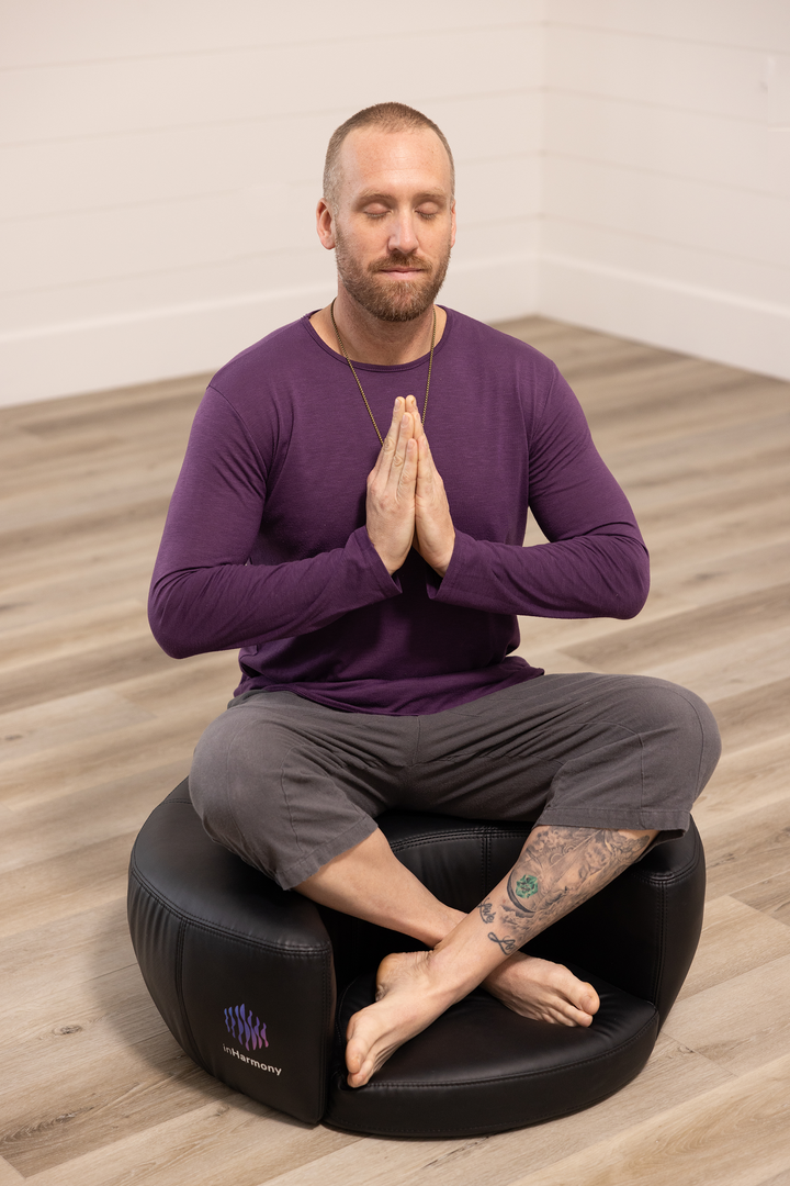 A man sitting atop a cushion in the middle of an empty room, cross legged, eyes closed, palms together in his chest