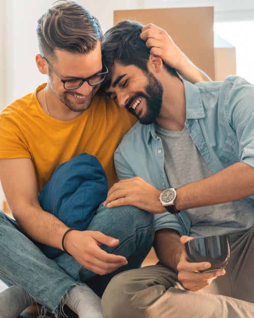 2 men as couple on indian-sitting position, laughing while one holding his head and the other touching the other's knee