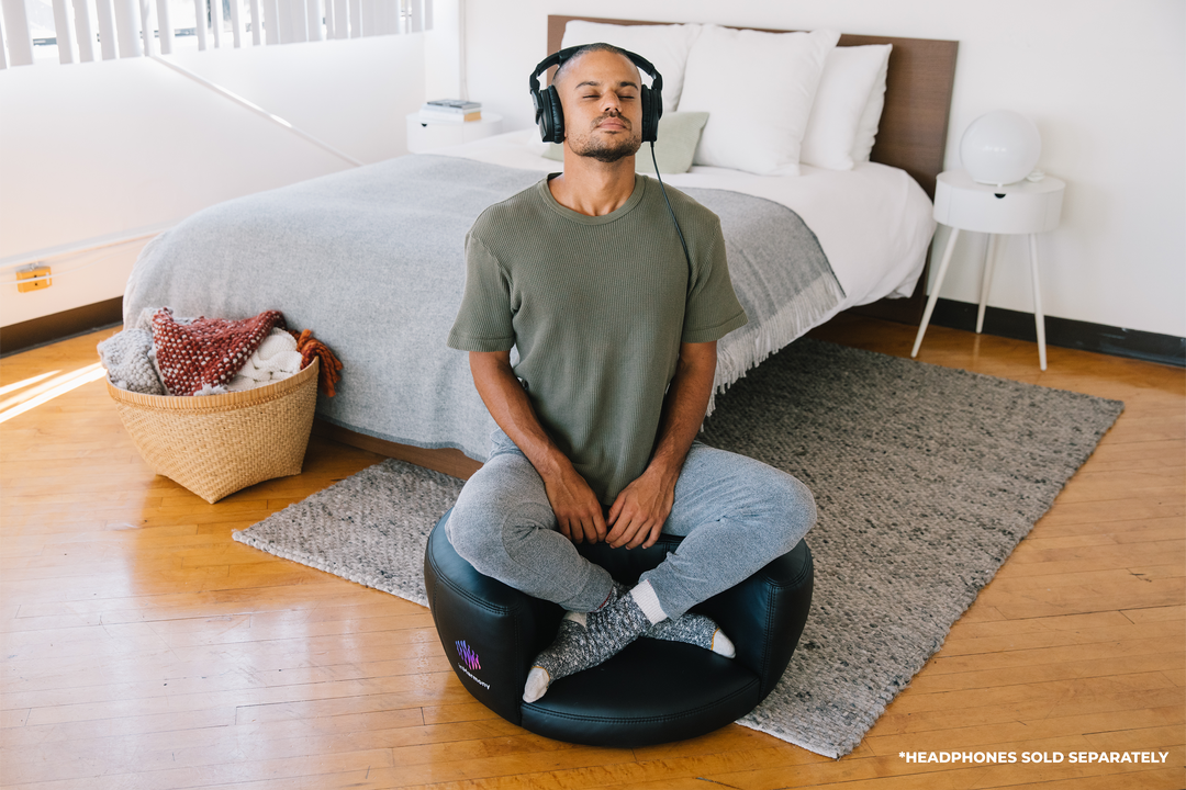 A man sitting atop a cushion, in the middle of a bedroom, his eyes closed with a headphone, cross legged in an elevated manner, his hands relaxed on his legs.