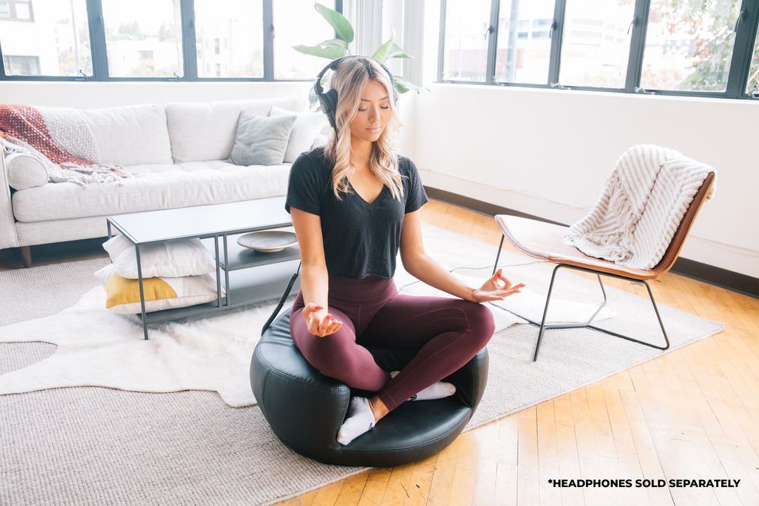 A girl in the middle of a living room, sitting atop a cushion where she is cross legged in an elevated manner, eyes closed, with a headphones over her head, hands on her knees, face up, her index and thumb touching.