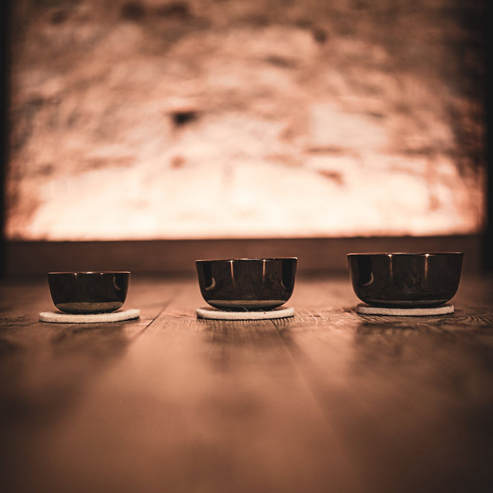 A leveled view of a three sized bowls, placed on the floor, with a cushion underneath them, with a background that's out of focus but seems to emanate light in a rocky wall