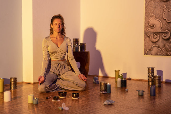 A woman sitting cross legged in the middle of room, palms resting on her knees, faced upwards, with her eyes closed. In front of her is a big bowl with a rod on its side. She is surrounded with candles and crystals laid on the floor.