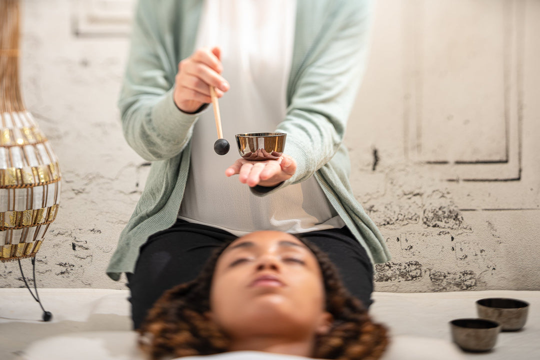 In the foreground is an out of focus head lying on the floor. While a person is seats halfway on her ankles above the lying person, where she holds a small bowl on her left hand, and her right hand holds the small, thin mallet