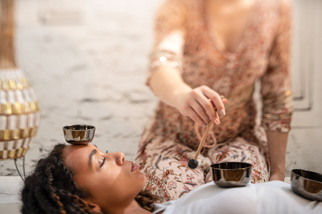 A woman is lying horizontally on the floor with bowls on her forehead, chest and stomach. Another woman is sitting on her knees towards her, holding a small wooden mallet, motioning to encircle it in the bowl in her chest