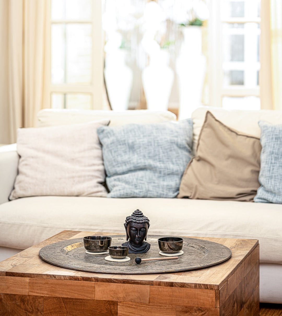 Three sized bowls with a cushion underneath and a thin, small mallet are placed in the center of a wooden table together with a decorative small, buddha head. A couch and an open door in the background.