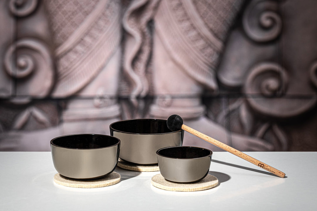 Three different sizes of bowls are placed on a flat surface with a cushion underneath and the small, thin mallet's head rests on the middle big bowl. A background of an out of focus feet that seems to be an ancient wall carving.