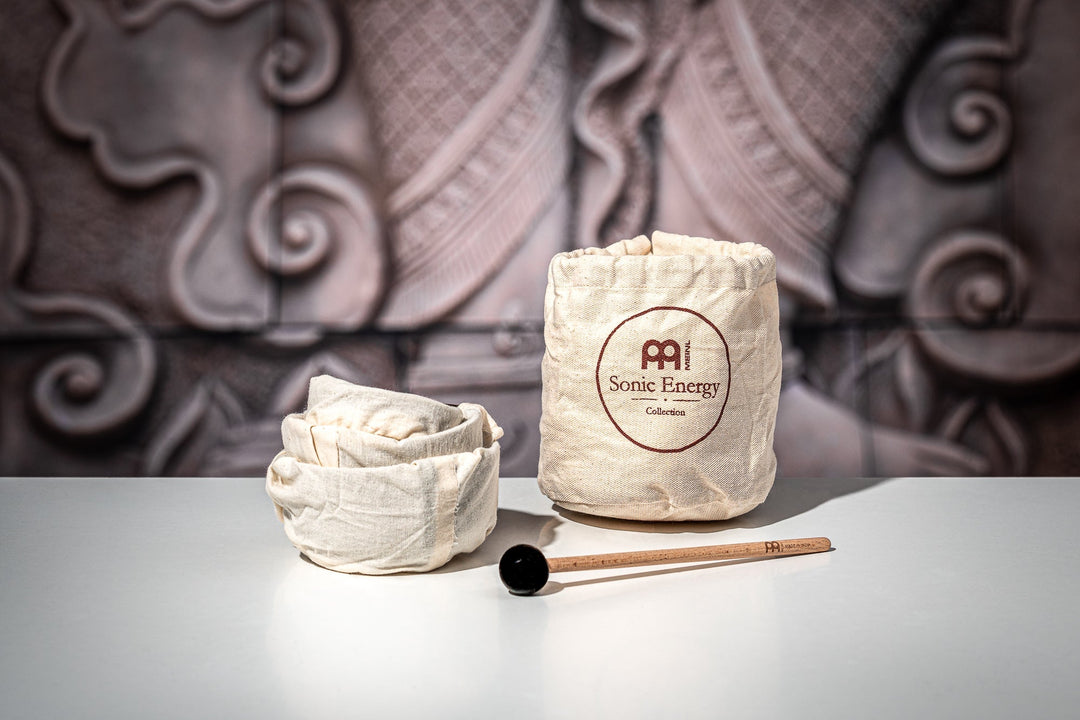 Three sized bowls are stacked together but with each inside their own clothing cover, on its right is a small bag for it. A small, thin mallet is placed fronting it.  In the background is an out of focus of what looks like an ancient stone carving of a feet.