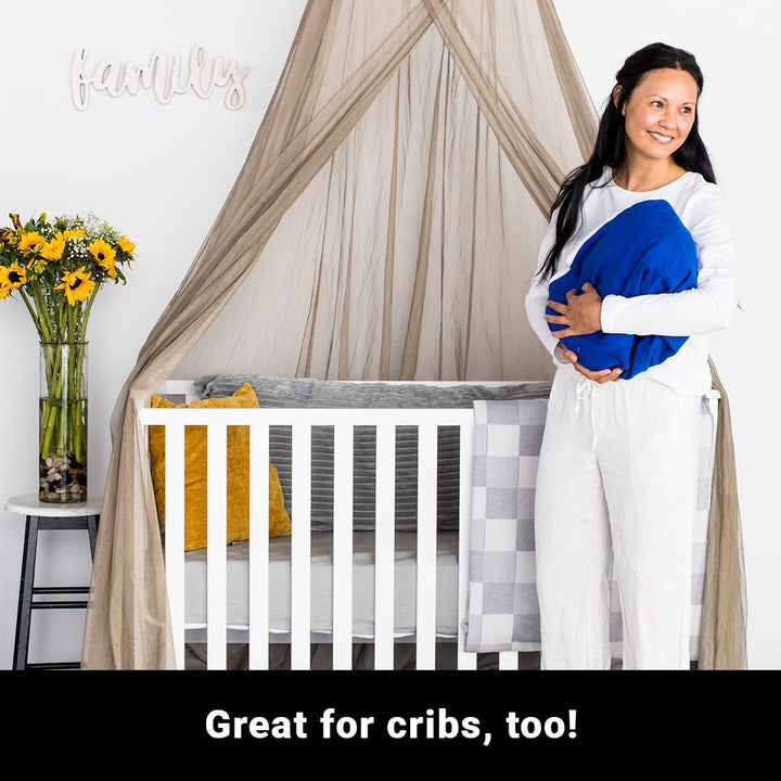 A woman, holding a ball of blanket which may be a child representation, standing in front of a crib where a fabric drapes over it from above. A vase with flowers is placed on top of a chair on the right side. A text below says, great for cribs, too.